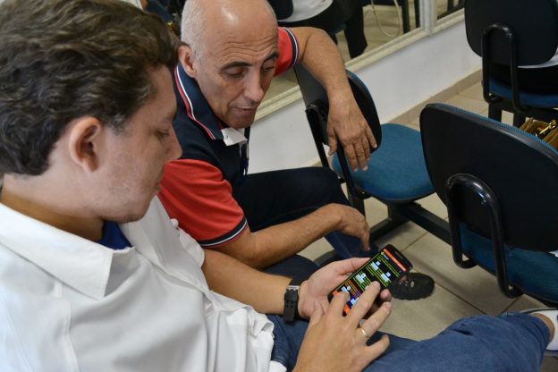 Descrição da imagem: foto de um homem manuseando um celular. Outro homem ao lado dele observa o aparelho.