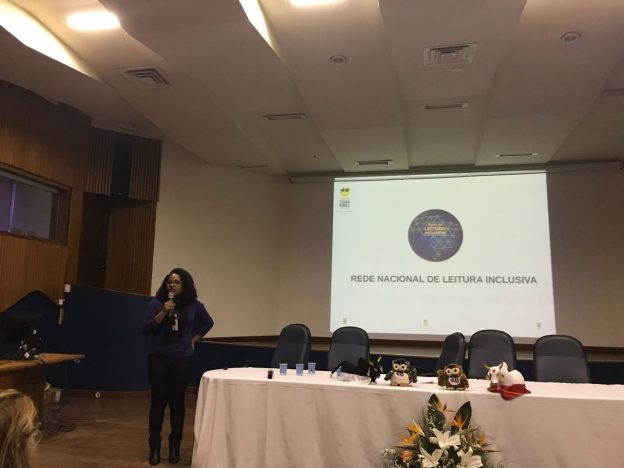 Descrição da imagem: foto de uma mulher falando ao microfone. Atrás dela, um telão com o logotipo da Rede Nacional de Leitura Inclusiva.