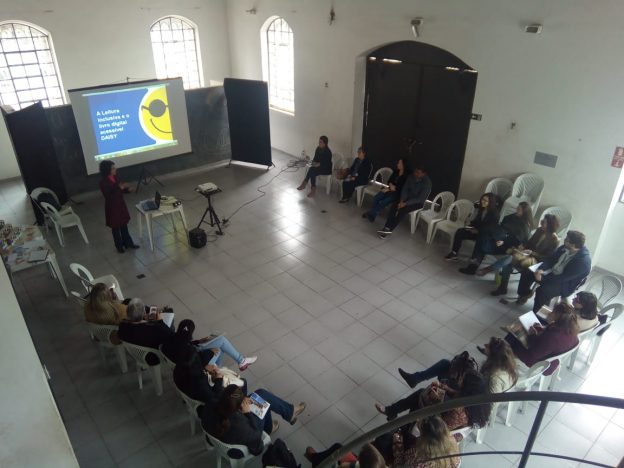 Descrição da imagem: foto de um grupo de 20 pessoas sentadas em um semicírculo. À frente deles há uma mulher de pé e um telão com o logotipo da Fundação Dorina.